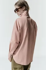 Camisa de cuello alto con cierre frontal cruzado mediante botones de carey. Presenta mangas largas con puños rectos y un diseño de bajo asimétrico con corte holgado.