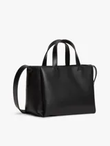 Cartera tote de poliuretano sintético liso color negro de la marca Tommy Jeans, con dos asas superiores y correa ajustable. Presenta cierre de cremallera, compartimento principal e insignia metálica de la marca en la parte delantera. Incluye un adorno colgante extraíble con cordón azul trenzado, dije en forma de corazón y herrajes plateados.