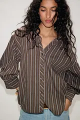 Camisa de cuello solapa con estampado de rayas verticales en tonos marrón y blanco. Presenta mangas abullonadas por debajo del codo con puños definidos y cierre frontal con botones.