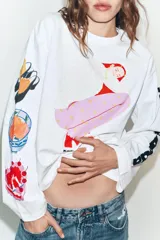 Camiseta blanca de corte oversize con cuello redondo y manga larga. Presenta una estampa gráfica central de una figura femenina, detalles estampados en la manga derecha y texto en la manga izquierda.