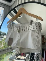 Short blanco de cuero sintético con cinturón con hebilla metálica y detalles de ojalillos metálicos. Presenta cordones cruzados en los laterales.