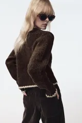 Chaqueta de punto de textura bouclé en color marrón oscuro, con cuello redondo y manga larga. Presenta detalles de vivo a contraste en color crema en los bordes y cierre frontal mediante lazadas decorativas con flecos.