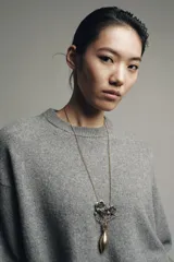 Collar largo con cadena de eslabones finos en tono dorado envejecido. El colgante consiste en un jarrón alargado dorado envejecido del cual emergen tres flores oscuras con detalles en relieve y hojas pequeñas.