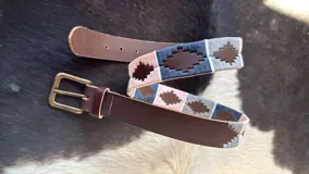 Cinturón de cuero marrón con detalles bordados a mano en hilos encerados con patrones geométricos en tonos azul, rosado y gris, hebilla metálica de bronce envejecido y terminaciones en cuero.