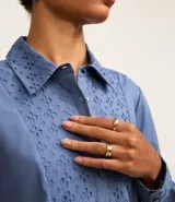 Vestido corto de corte evasé confeccionado en tricolina de algodón, color azul. Presenta cuello camisero, cierre frontal de botones y un panel central con detalle bordado calado. Posee mangas largas con puños abotonados.