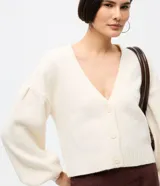 Cardigan de punto color crema con escote en V, cierre frontal de botones y mangas largas abullonadas con puños acanalados. Presenta un diseño corto y ajuste cómodo.