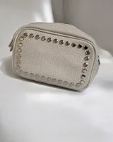 Cartera bandolera de cuero sintético color beige, con diseño rectangular y detalle de tachas metálicas circulares en el contorno frontal. Cuenta con correa larga ajustable y desmontable mediante mosquetones, cierre superior y bolsillos internos y externos.