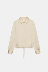 Camisa de cuello solapa y manga larga con puños anchos, confeccionada en tejido satinado color beige. Presenta cierre frontal con botones ocultos y bajo ajustable mediante cordones del mismo tejido.