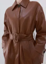 Chaqueta larga de cuero sintético marrón con efecto charol, solapas clásicas, cierre frontal oculto y cinturón ajustable con hebilla del mismo material. Cuenta con bolsillos laterales y mangas largas con trabillas en los puños.