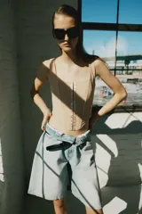Top corsetero de tul semitransparente color beige, con escote cuadrado, tirantes anchos y cierre frontal con ganchos metálicos. Presenta bajo asimétrico y costuras reforzadas con ribetes a tono.