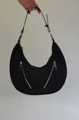 Cartera mediana tipo hobo de cuero sintético color gris, con diseño semicircular, dos bolsillos externos frontales con cierre y correa de hombro ajustable.