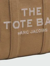 Bolso tipo tote Marc Jacobs modelo The Small Tote, color beige con detalles y texto en negro que dice "THE TOTE BAG MARC JACOBS". Cuenta con asas superiores y laterales para llevar en la mano o al hombro.