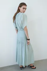Vestido midi de tejido texturizado color verde menta, con escote redondo fruncido, espalda semiabierta con breteles finos y capa decorativa sobre los hombros. Cuenta con sección de punto smock en la cadera y falda con leve vuelo.