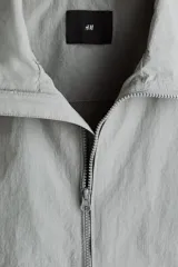 Campera rompevientos de nylon color gris, repelente al agua, con cuello elevado, cierre frontal y bolsillos laterales con botón a presión oculto.