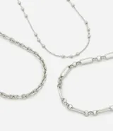 Set de tres collares de metal plateado con diferentes texturas: una cadena de eslabones pequeños, una cadena de eslabones alargados y una cadena fina con detalles de esferas.