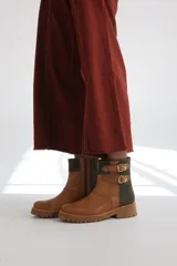 Bota estilo biker de caña media, confeccionada en cuero negro con detalles en gamuza marrón. Presenta correas ajustables con hebillas metálicas, tachas decorativas y suela track dentada.