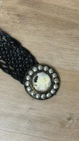 Cinto de cuero trenzado color negro con hebilla circular grande de metal plateado, decorada con tachas esféricas y centro de pelo natural con manchas.