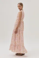 Vestido largo de algodón color rosa con estampado floral en tonos beige y dorado. Tiene cuello alto con lazo ajustable, corte imperio, falda escalonada con volados y diseño sin mangas.