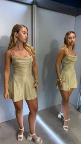 Vestido corto de fiesta con acabado brillante y efecto glitter. Presenta un escote drapeado tipo cowl neck, tirantes finos y una falda con corte evasé que aporta volumen y movimiento.