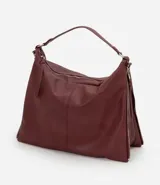 Bolso tipo hobo de gran tamaño en color bordó, confeccionado en poliuretano. Presenta una estructura desestructurada, cierre superior, correas dobles (una de mano y una larga desmontable) y cierres laterales decorativos con tiradores.
