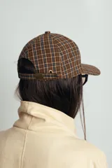 Gorra tipo baseball con estampado de cuadros escoceses en tonos azul, naranja y beige. Presenta un parche de cuero sintético en el frente y cierre ajustable en la parte posterior.