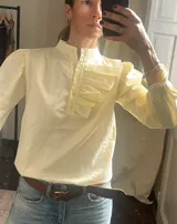 Camisa de manga larga color amarillo claro con cuello alto mao, cierre frontal con botones y detalle de volados plisados en el pecho. Presenta mangas abullonadas con hombros fruncidos.