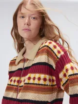 Campera de punto estampada de corte holgado, con cierre metálico frontal y cuello de corderito sintético en color tostado. Presenta un diseño de franjas horizontales con patrones geométricos en tonos bordó, mostaza, marrón y beige.