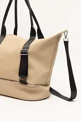 Bolso tipo tote color beige con detalles en negro, confeccionado en material sintético liso. Cuenta con doble asa superior, correa ajustable y extraíble, cierre principal superior y un compartimento inferior independiente con cierre. Presenta correas decorativas verticales con hebillas metálicas y logo grabado en la base.