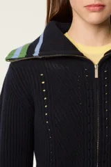 Cárdigan de punto azul marino con cierre frontal de cremallera. Presenta cuello tipo camionero y puños con detalles de rayas en tonos verde y celeste. El tejido tiene una textura acanalada con detalles calados decorativos.