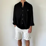 Camisa de lino blanca con cuello mao y cierre frontal de botones. Presenta un pequeño bordado oscuro en el pecho y mangas largas con puños doblados.