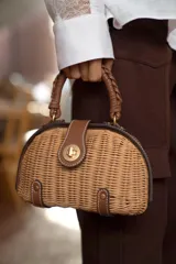 Bolso rígido tipo bandolera de mimbre trenzado en tono natural, con detalles en cuero sintético marrón, asa superior trenzada y cierre metálico dorado.
