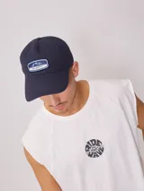 Gorro de visera estilo dad hat, color azul marino, confeccionado en lona lavada. Presenta un parche rectangular bordado en el frente con el logo de la marca Rusty.