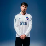 Camiseta de fútbol Umbro del Club Nacional de Football, modelo Home de manga larga. Diseño en color blanco con detalles en azul y rojo en el cuello y los puños. Incluye el logo de Umbro, el escudo del club y el patrocinador Antel en el frente.
