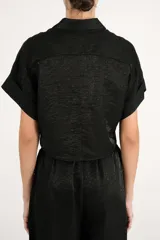 Camisa crop negra de tejido fluido y acabado brillante, con cuello camisero, mangas cortas con vuelta y nudo frontal en el dobladillo.