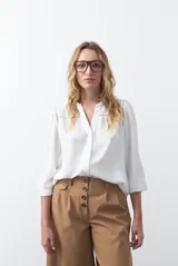 Camisa blanca de corte holgado con escote en V, cierre frontal de botones y mangas tres cuartos. Presenta detalles de encaje calado en los hombros y canesú, con una textura sutil en la tela.