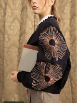 Campera de jogging negra con cierre frontal, cuello con detalle de volados y bordados florales en las mangas. Presenta un corte holgado y puños elásticos.