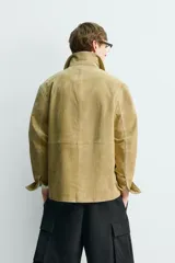 Cazadora de hombre de corte relajado, confeccionada en piel con acabado ante color mostaza/verde oliva. Presenta cuello solapa, manga larga con puño abotonado, cierre frontal de botonadura y cuatro bolsillos de plastrón visibles (uno en el pecho y tres en la parte inferior).