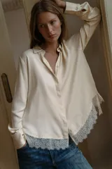 Camisa de satén color crema con cuello solapa, manga larga y cierre frontal con botones ocultos. Presenta un detalle de encaje en el bajo asimétrico.