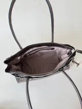 Cartera de cuero de tamaño medio con estructura rígida. Presenta una tira decorativa con tachas y hebillas metálicas en los extremos, además de lazos laterales. Cuenta con doble asa para llevar al hombro y cierre superior.