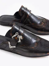 Mocasín tipo slide color marrón oscuro con efecto gastado, confeccionado en cuero vacuno. Presenta detalles de picado en la capellada y laterales, forrería de cuero vacuno, hebilla y puntera metálica.