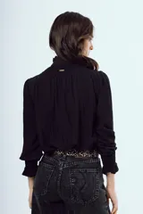 Camisa negra de manga larga con cuello alto gorguera y detalles fruncidos con nido de abeja en el pecho y los puños. Cierre frontal con botones.