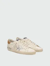 Zapatillas Golden Goose modelo Super-Star de cuero napa color blanco, con la icónica estrella lateral laminada en plateado, talón de gamuza marrón y efecto desgastado artesanal. Presenta inscripciones y detalles escritos a mano en los laterales y la lengüeta.