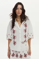 Blusa blanca de gasa de algodón con mangas 3/4 abullonadas y escote en V con botones. Cuenta con bordados en tonos marrones y naranjas en la zona frontal y las mangas.