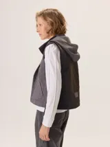 Chaleco gris con cuello alto y capucha, cierre frontal con botones a presión. La parte delantera es de lana gris lisa y la parte interior del cuello y el forro son de tela con patrón de espiga en blanco y negro.