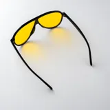 Lentes de sol con armazón de policarbonato negro y diseño de estilo aviador con puente superior plano. Las lentes son de color amarillo y cuentan con filtro UV400. Las patillas incorporan detalles metálicos dorados en las bisagras.