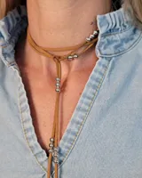 Collar estilo corbatín confeccionado en tiento de cuero color suela, con detalle de cuentas metálicas plateadas distribuidas a lo largo de las tiras.