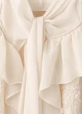 Blusa color marfil de tejido fluido, con cuello alto con lazo, volados frontales y mangas largas acampanadas con detalles florales bordados.