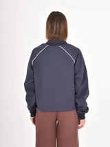 Buzo azul oscuro de corte holgado con cuello polo y escote en V. Presenta mangas largas con costuras raglán en color blanco y un estampado frontal con la palabra Ambience y el texto United Nation League en tonos dorados.