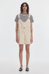 Monoprenda corta de denim color beige, de fit oversize, con tirantes regulables y bolsillo canguro en el frente.