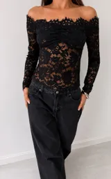 Top de encaje negro con diseño off-shoulder, mangas largas y detalle fruncido en el centro del busto.